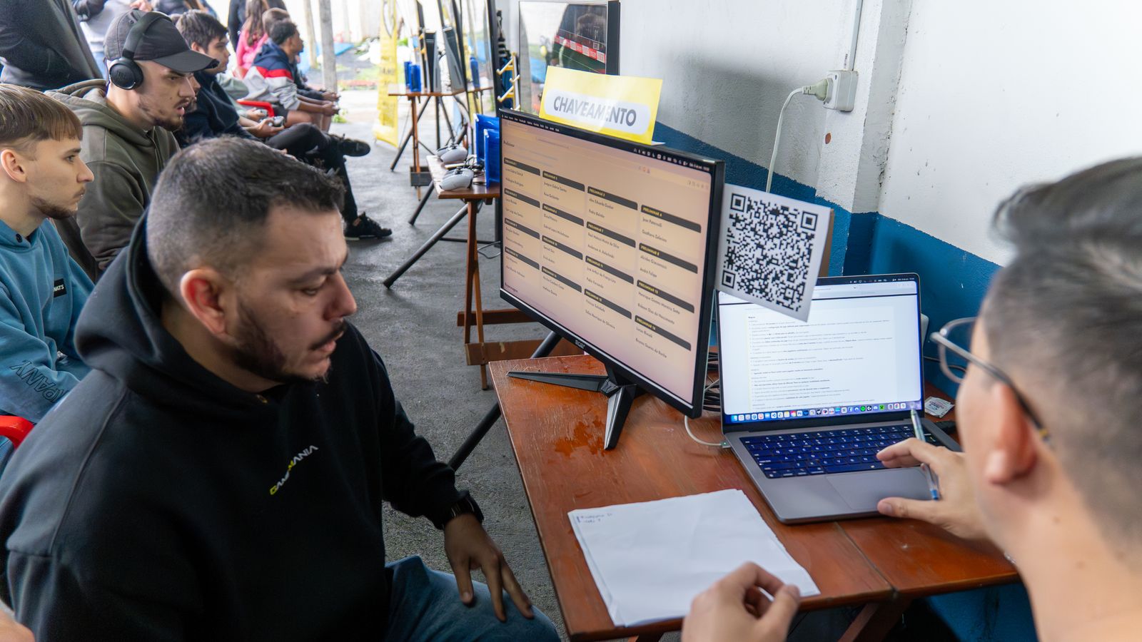 Equipe Game Mania atualizando o chaveamento do campeonato em um monitor, com QR Code de acesso ao lado